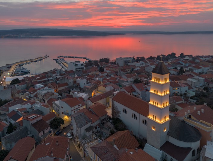Aerial view of Crikvenica at sunset, featuring a lit church tower and the Adriatic Sea in the background.