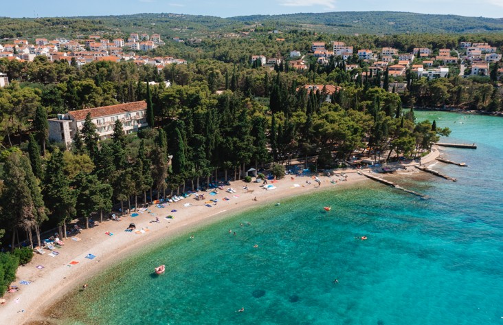 Aerial view of a pebble beach and turquoise bay surrounded by pine trees at Aminess Vival Velaris Resort.
