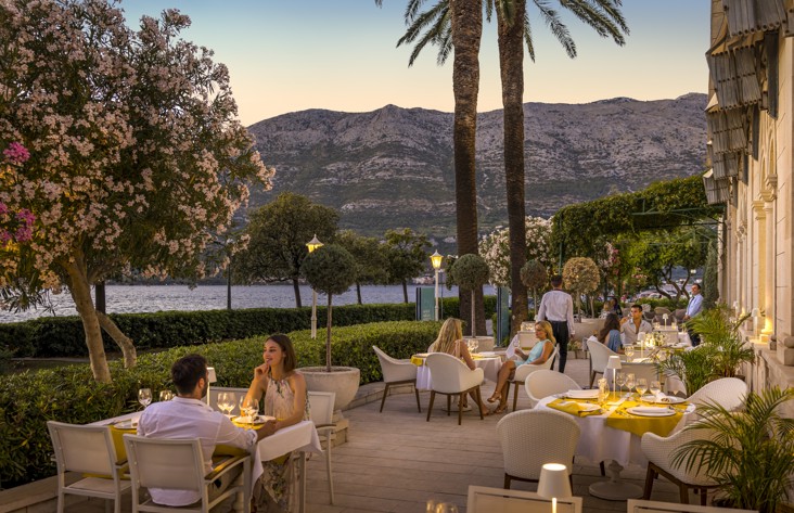 Gosti uživaju u romantičnoj večeri na terasi restorana i bara 7 Seas u Aminess Korcula Heritage Hotelu za vrijeme zalaska sunca, okruženi cvatućim oleandrima, palmama i pogledom na Pelješke brežuljke preko sjajnog mora.