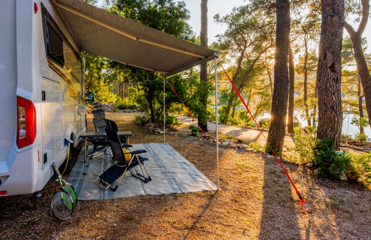 Ein Auto-Campingplatz bei Aminess Port9 Planet Camping Korcula, mit einem Wohnmobil geparkt unter Kiefern, einem schattigen Sitzbereich und Blick auf die umliegende Naturlandschaft