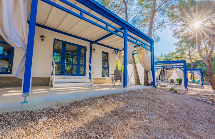 Exterieur van vakantiehuizen bij Port9 Planet Camping door Aminess, holiday homes, met blauwe kozijnen, schaduwrijke terrassen, en omgeven door pijnbomen met zonlicht dat erdoorheen filtert.