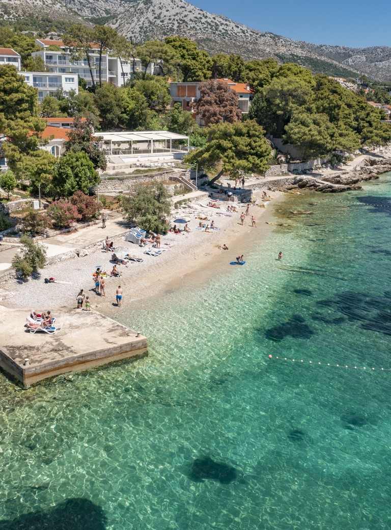 Pogled iz zraka na plažu blizu Aminess Maradiso Casa Bellevue, prikazuje kristalno čisto tirkizno more in živahno zelenje.