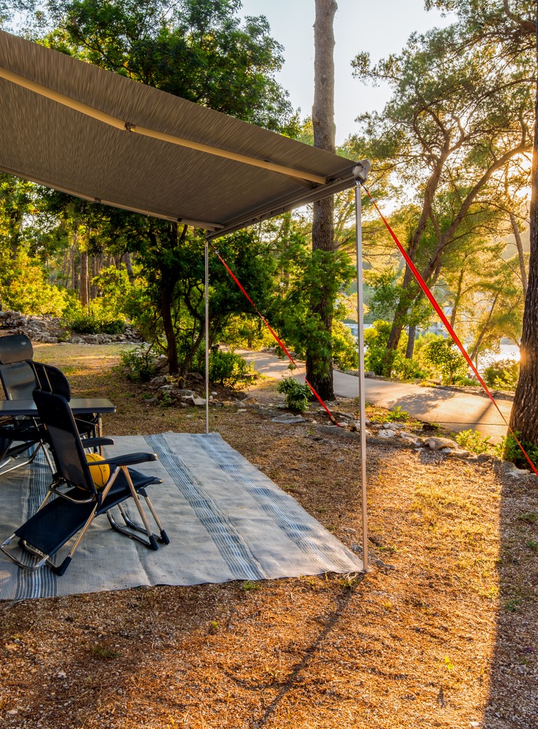 Un sito di campeggio auto presso Aminess Port9 Planet Camping Korcula, con un camper parcheggiato sotto gli alberi di pino con un'area salotto ombreggiata e una vista sul paesaggio naturale circostante