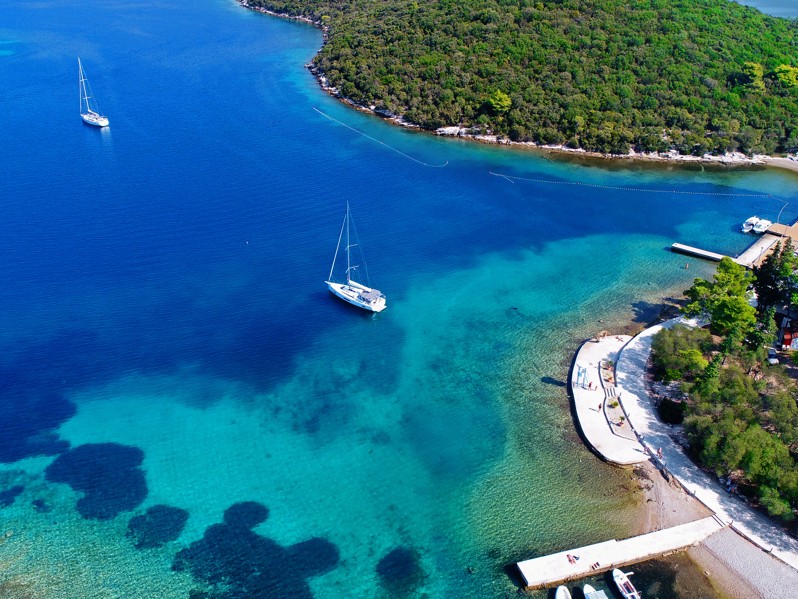 Pogled iz zraka na uvalu kod Port9 Planet Camping by Aminess, Korčula, prikazuje živopisne tirkizne vode, maleni otok, usidrene brodove i bujne zelene borove šume koje okružuju kamp, pod vedrim plavim nebom.