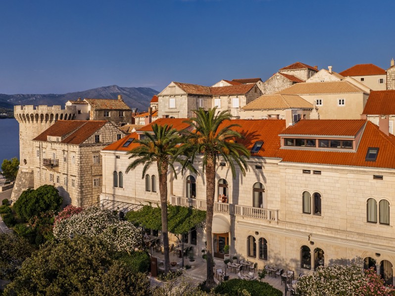 Historic stone Aminess hotel by the sea with palm trees and red roofs.