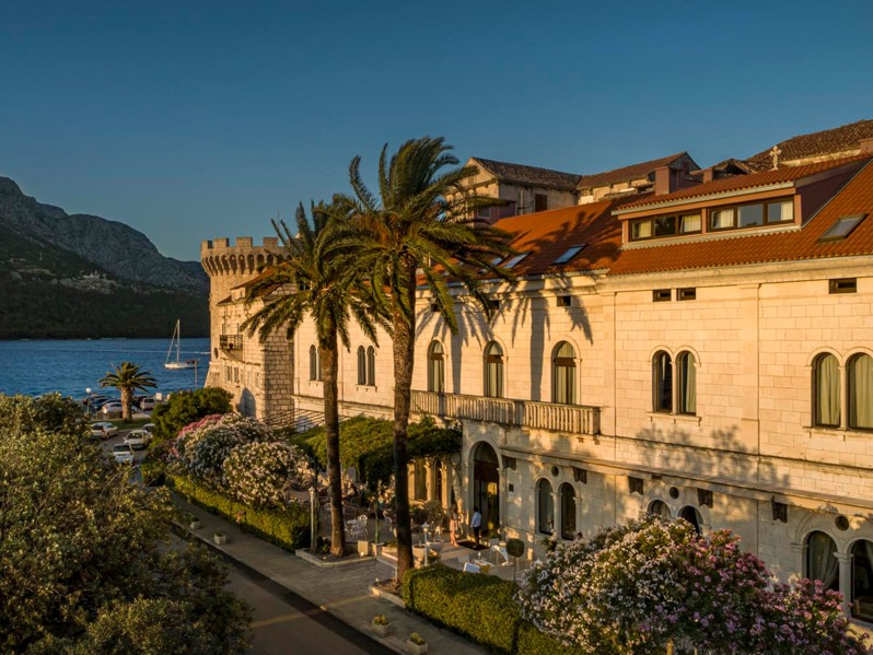 Aminess Younique Korcula Heritage Hotel, povijesna zgrada s crvenim krovovima u starom dijelu Korčule blizu mora, smještena pored Kule Kanalevic, okružena palmama i cvatućim vrtovima, snimljena tijekom zlatnog sata.