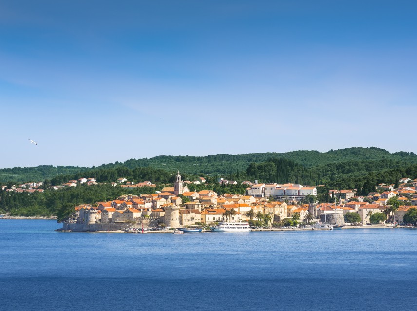 Korcula Old Town on Korcula Island with terracotta rooftops, green hills, and the Adriatic Sea under a clear blue sky.