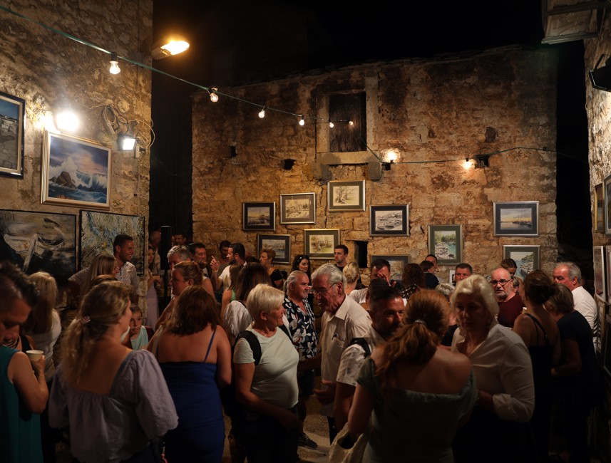 A crowd of people attending an exhibition of paintings and photographs on Brač Island, Croatia, showcasing local art and culture.