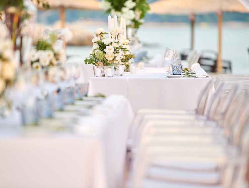 The image shows a wedding setup with details of flowers and candles inside a tent, transparent chairs, and the sea in the background.