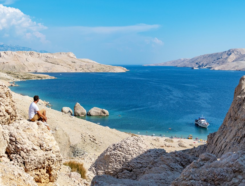 Un uomo siede sulla costa rocciosa di Pag con vista sul mare blu e una barca.