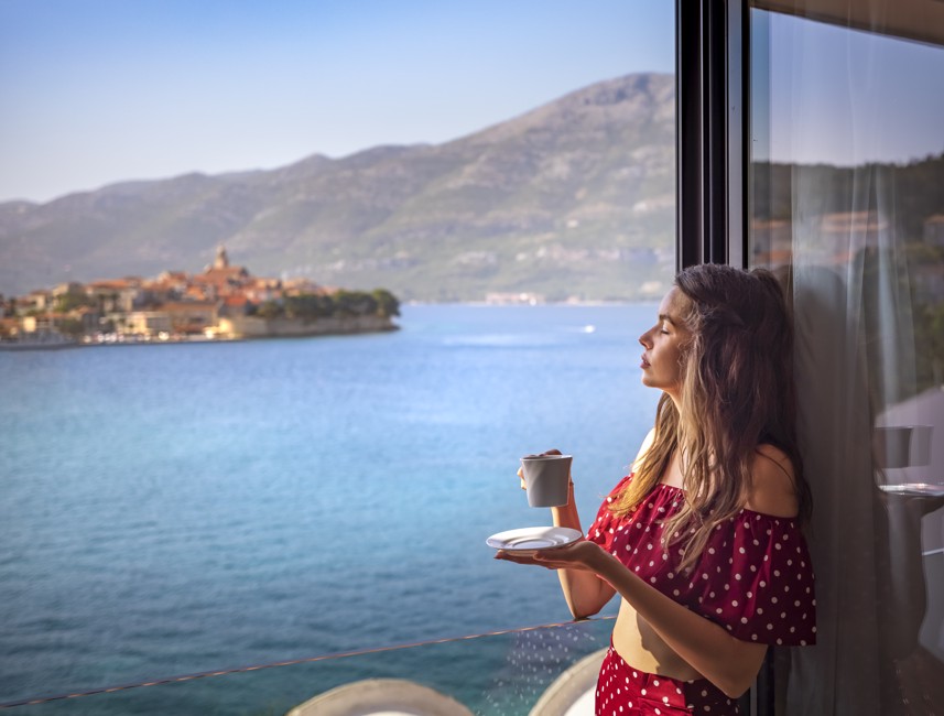 Frau genießt ihren Morgenkaffee auf dem Balkon eines Aminess Zimmers mit ruhigem Meerblick und Küstenort