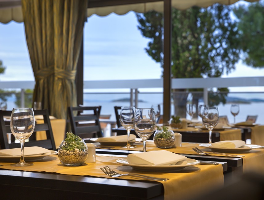 Dining tables set on a terrace with a beautiful view of the sea and greenery at Aminess Vival Grand Azur Hotel.