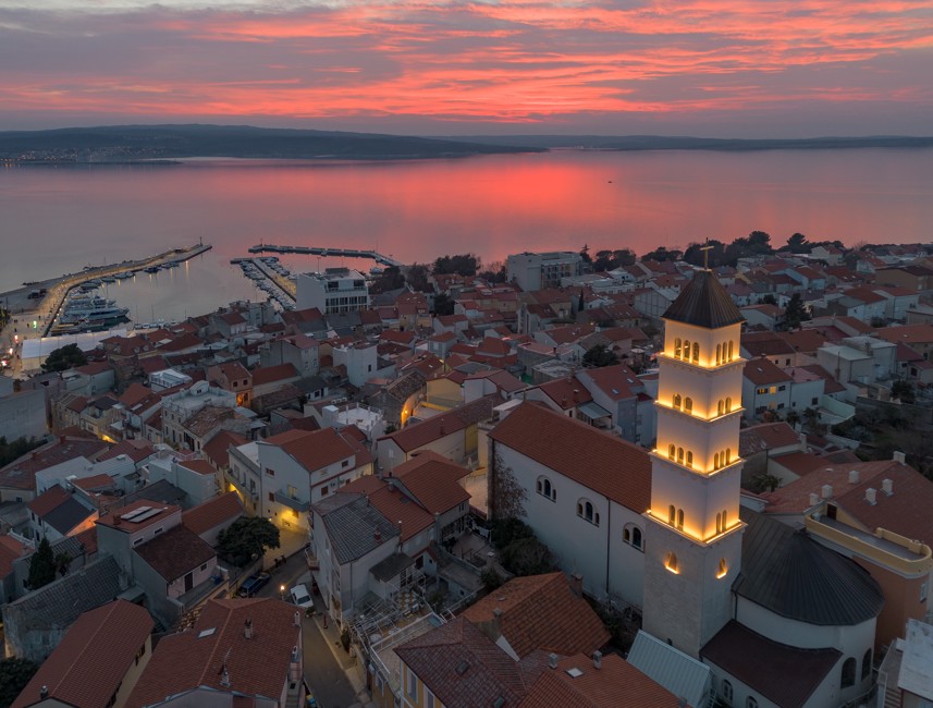 Aerial view of Crikvenica at sunset, featuring a lit church tower and the Adriatic Sea in the background.