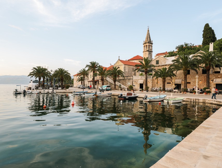 A small bay in the city of Brač Island, Croatia, with fishing boats docked along the shore, a church and bell tower in the background, and palm trees lining the promenade.