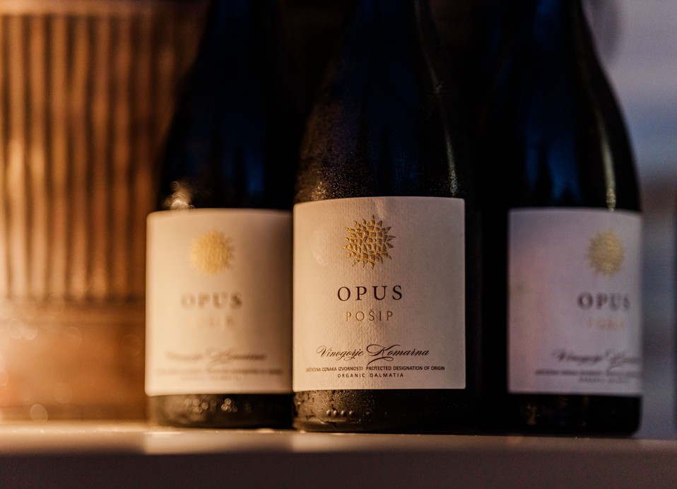 Butelki wina Opus Posip z Vinogorje Komarna są prezentowane w ciepłym świetle.