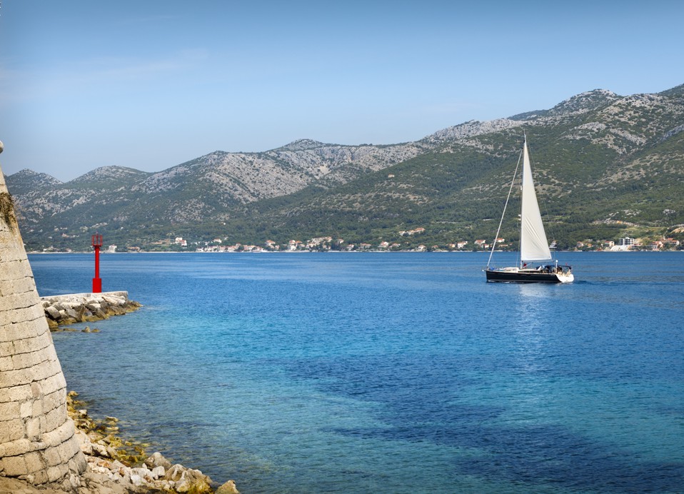 Een zeilboot glijdt langs de Puntin-toren in Korčula, met de historische stenen structuur op de waterkant, omlijst door kristalhelder Adriatisch water
