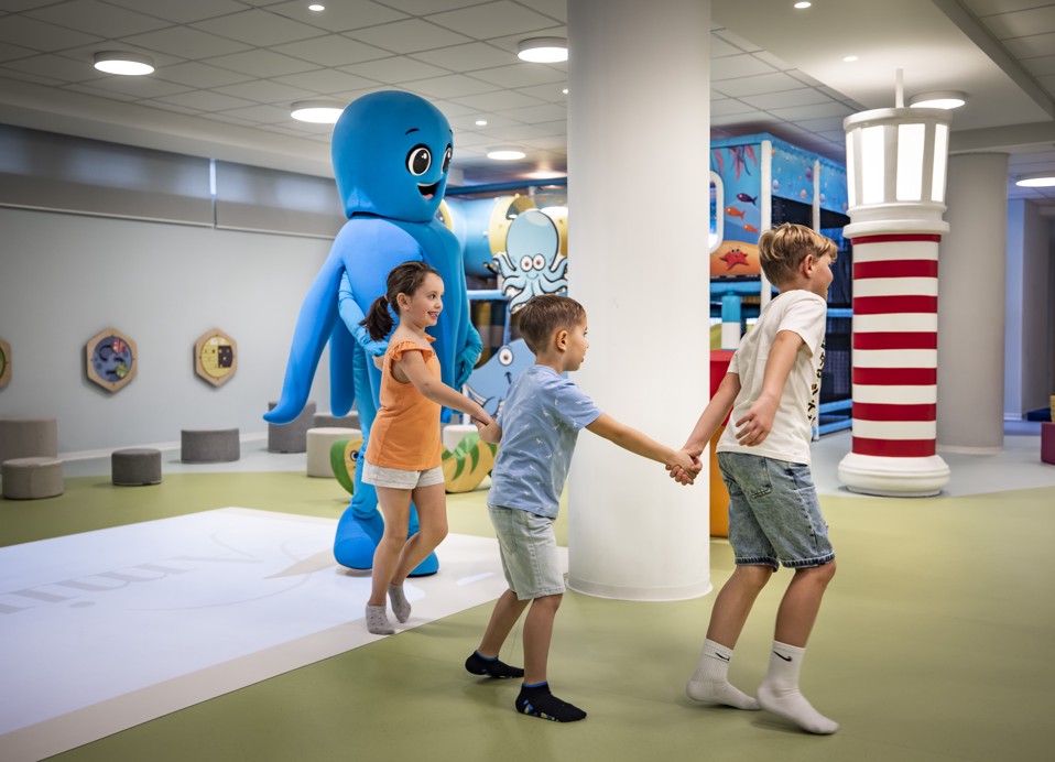 Bambini che si tengono per mano e giocano con la mascotte Mirami nella sala giochi.