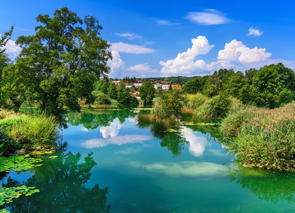 The green Mrežnica river with lush banks and a village behind, author Aleksandar Gospić.