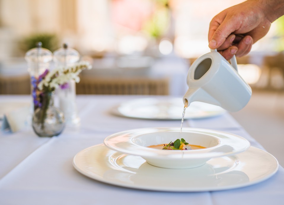 Cameriere che finisce elegantemente un piatto di zuppa al tavolo, versando un ricco brodo da una elegante brocca bianca su un piatto splendidamente presentato, migliorando l'esperienza culinaria con un tocco di sofisticazione presso Aminess Younique Korcu