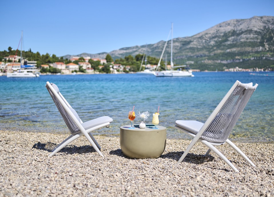 Opustajući ambijent u Puntomare Fun Beach zoni blizu Aminess Port9 Planet Camping Korčula, s prikazom stola na plaži s pićima, ležaljkama za sunčanje i slikovitim pogledom na obalu