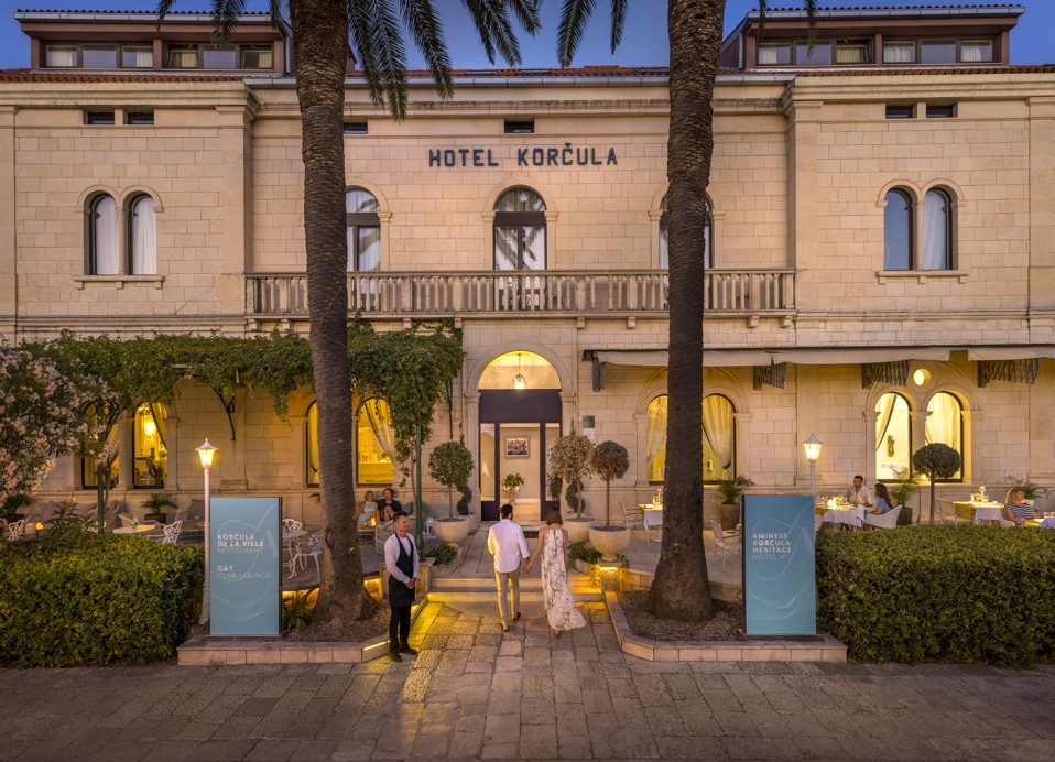 Fasada Aminess Younique Korcula Heritage Hotela u starom gradu Korčuli, osvijetljena toplim večernjim svjetlima, s parom koji ulazi u hotel i gostima koji večeraju na terasi okruženoj palmama.