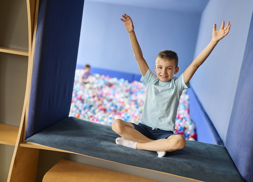 Een jongen zit in de speelruimte met uitzicht op het ballenbad en steekt blij zijn armen omhoog