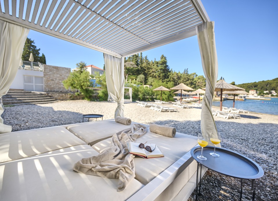 Ležalniki pod slamnatimi senčniki na prodnati plaži pri Port9 Planet Camping by Aminess, Korčula, z razgledom na morje in hribe.