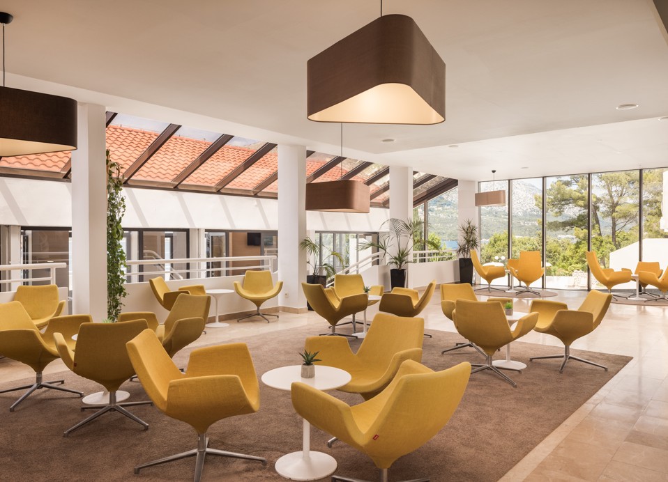 Un moderno atrio di hotel con tetto in vetro, sedie gialle e un bar con bottiglie e bicchieri in mostra a Korcula presso l'Aminess Younique Liburna Hotel.