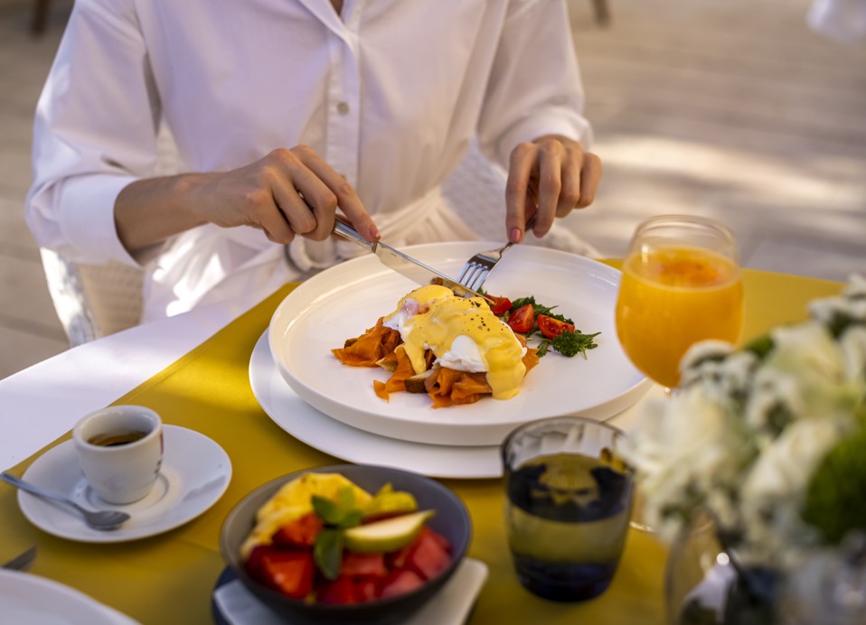 Persona in camicia di lino che si gode la colazione con Eggs Benedict, succo d'arancia e frutta sulla terrazza del 7 Seas Restaurant and Bar.