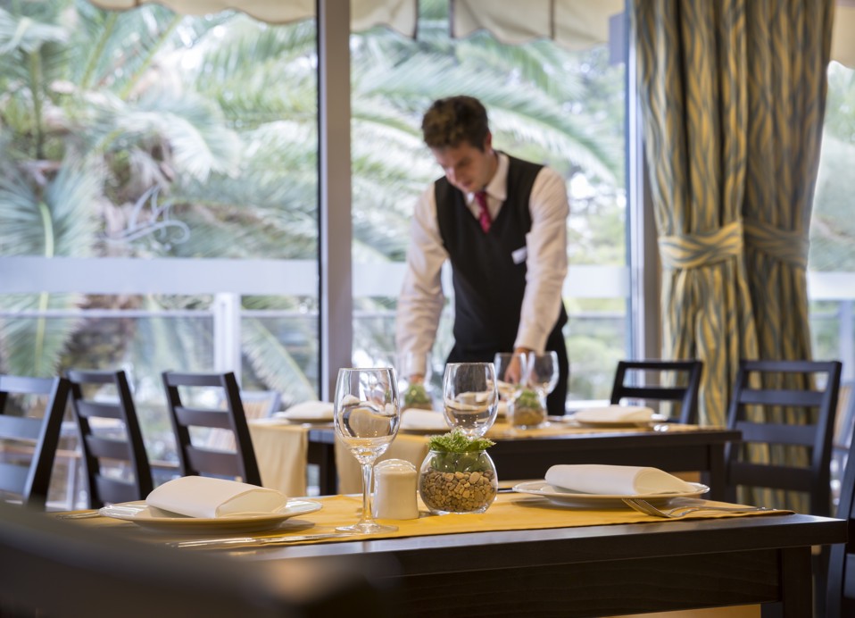 Ein Kellner deckt einen Esstisch in der Nähe großer Fenster in einem Buffet-Restaurant mit üppigem Grün draußen im Aminess Vival Grand Azur Hotel.