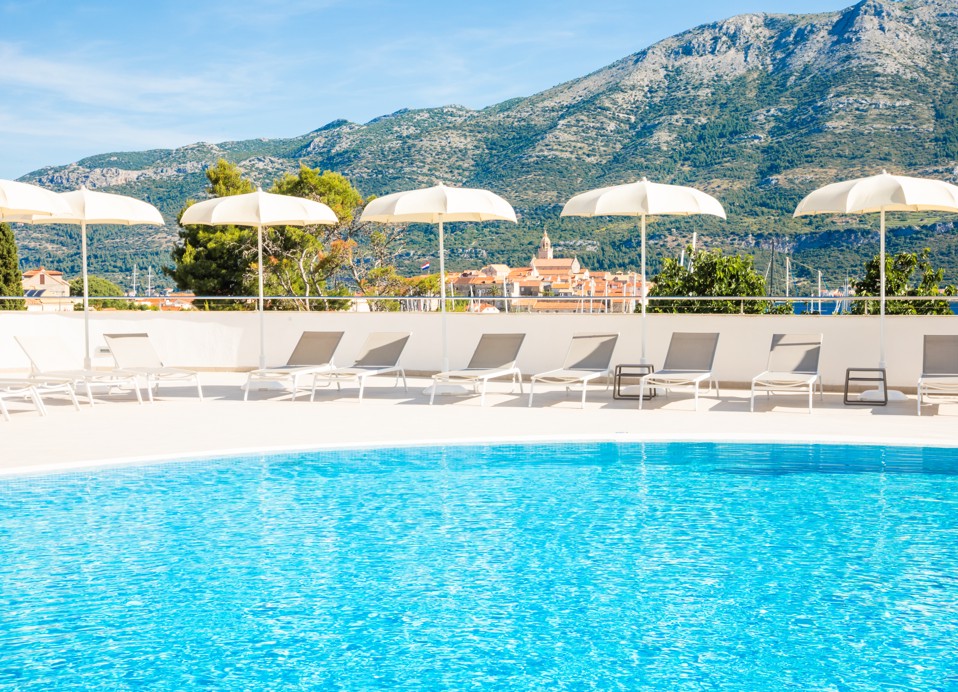 Outdoor pool area at Maradiso Marko Polo by Aminess in Korcula with sun loungers, umbrellas, and stunning views of surrounding hills.