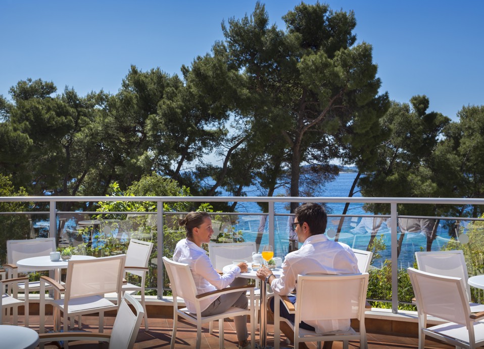Paar genießt Aperitif-Bar mit moderner weißer Sitzgelegenheit mit Blick auf die Pinien und das Meer im Aminess Vival Grand Azur Hotel.