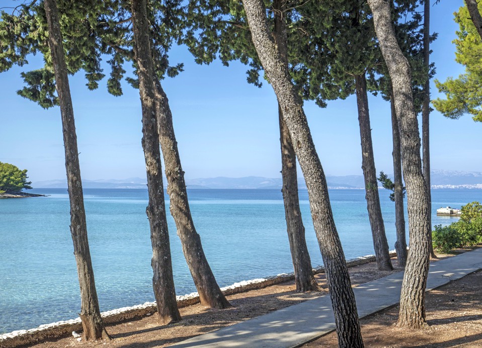 Obmorska promenada, obdana z borovci in kristalno čistim morjem pred Aminess Vival Velaris Resortom na otoku Braču, popolno mesto za sprostitev med poletnimi počitnicami na Hrvaškem.