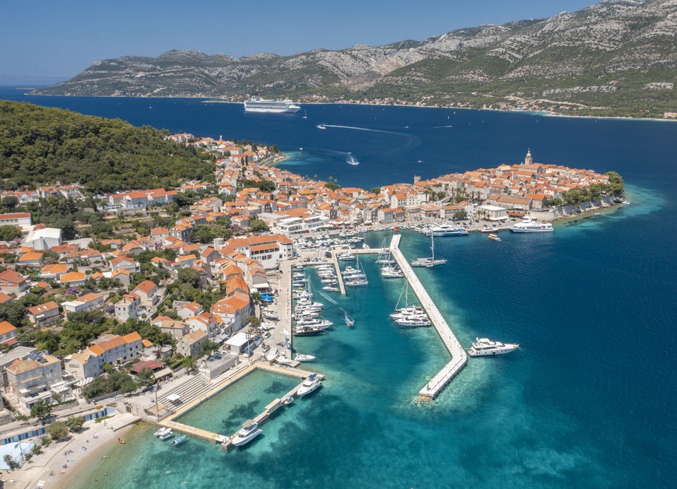 A breathtaking aerial view of Korcula town and its coastline, showcasing turquoise waters, a charming harbor, and terracotta-roofed buildings near the Maradiso Marko Polo by Aminess.