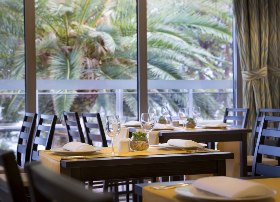 Innenansicht eines Restaurants mit gedeckten Tischen in der Nähe großer Fenster mit Blick auf grüne Palmen im Aminess Vival Grand Azur Hotel.