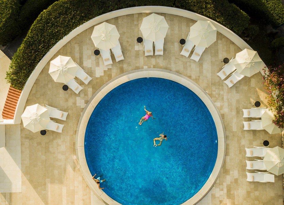 An aerial view of a pool with people swimming featuring sunbathing and relaxing loungers under umbrellas at Aminess Younique Liburna Hotel.