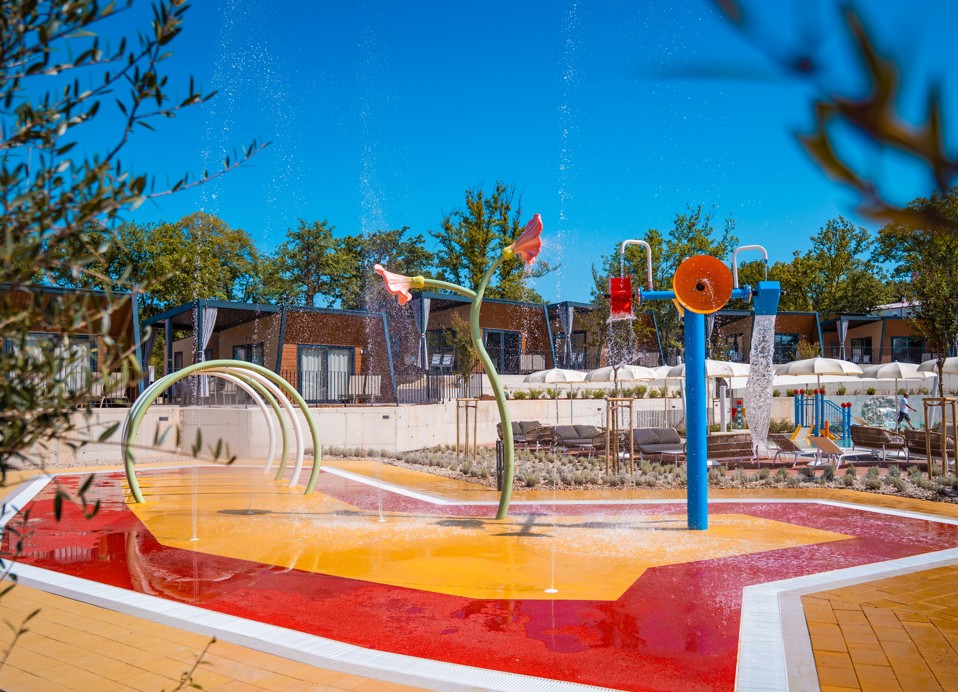 Parco giochi acquatico colorato con elementi interattivi e spruzzi d'acqua, circondato da case mobili presso l'Aminess Style Camping Atea Resort.