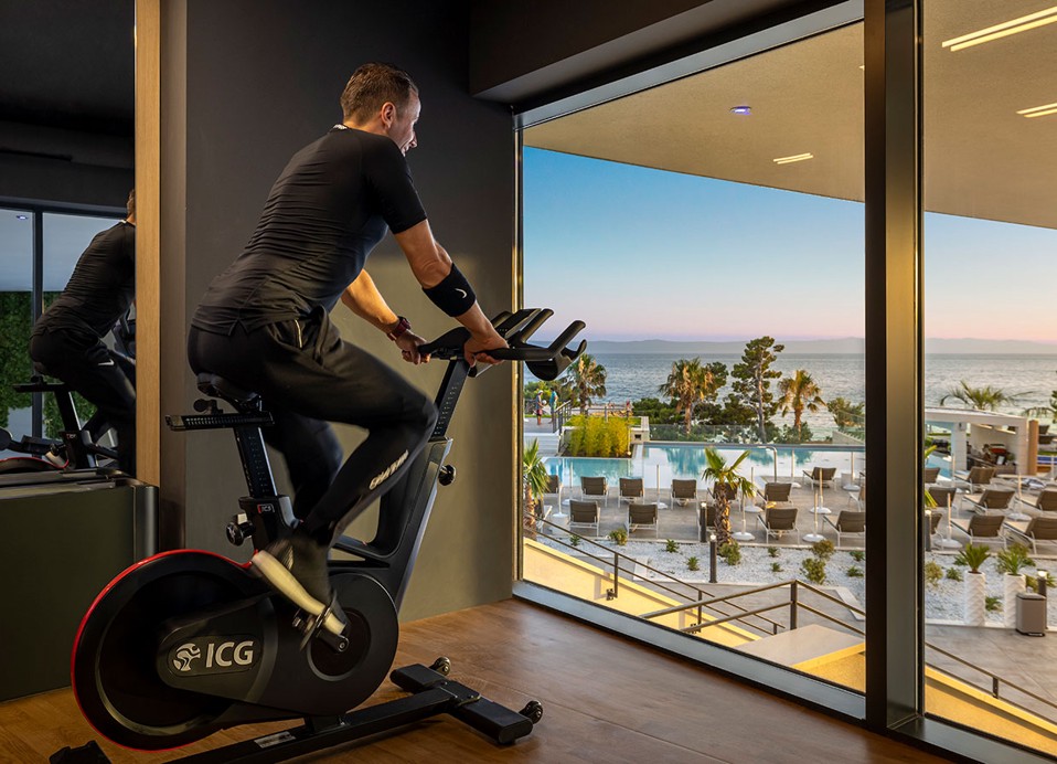 Un uomo pedala su una bicicletta in una palestra con vista sulla piscina interna presso l'hotel Aminess Laurel Khalani.