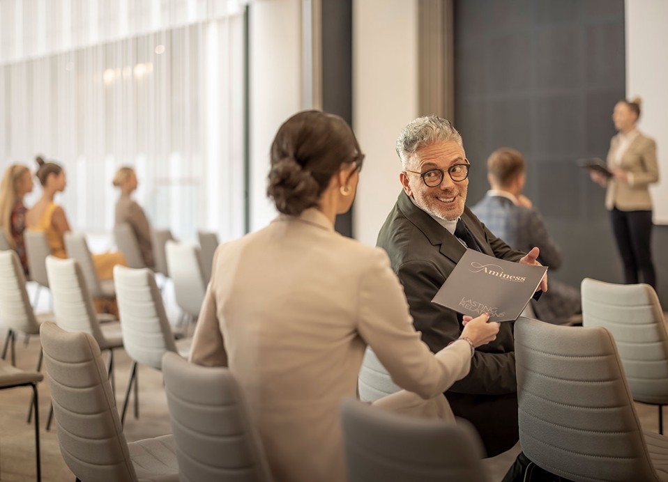A woman passes a folder to a man in a convention hall at Aminess Laurel Khalani hotel.