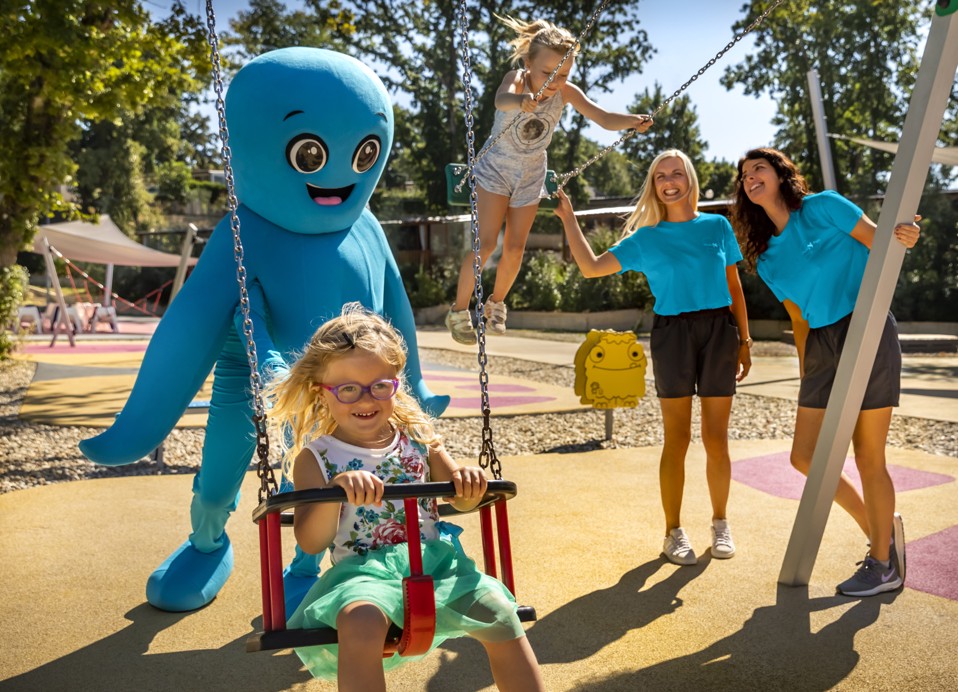 Kinder spielen auf Schaukeln und Rutschen mit dem Mirami Maskottchen und freundlichem Personal im Aminess Style Camping Atea Resort.