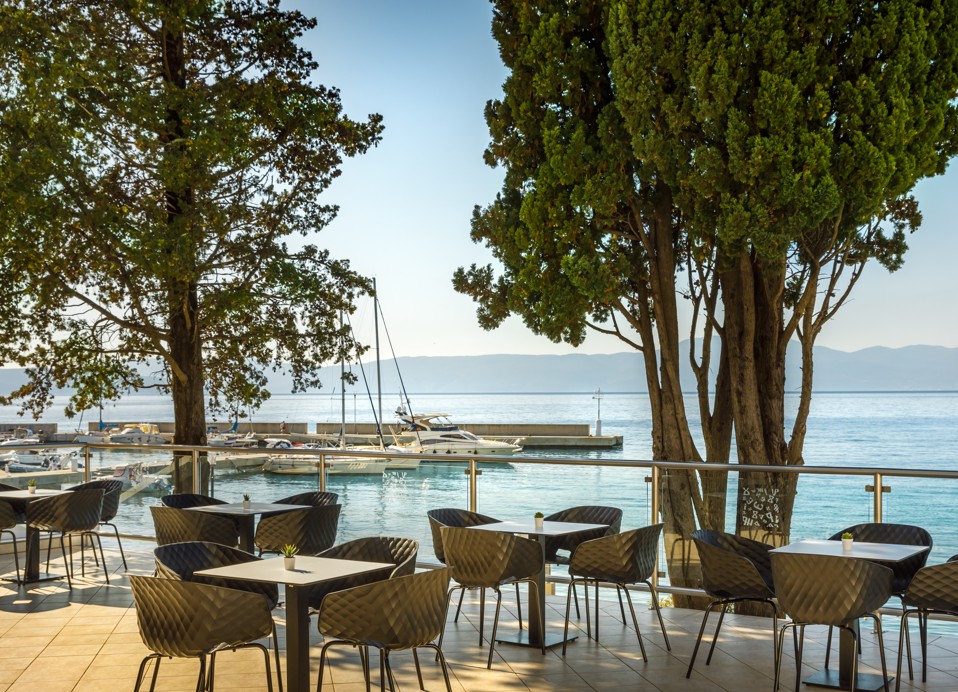 Sedežna površina Veya Lounge Bar z ratanastimi stoli in mizami, postavljenimi pod drevesa, s čudovitim pogledom na Jadransko morje v bližini Aminess Style Camping Atea Resort.