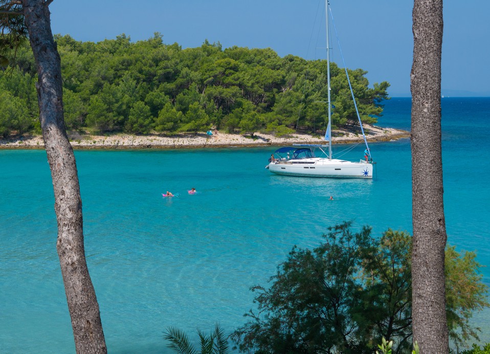 Miren zaliv s turkiznim morjem in jadrnico pred Aminess Vival Velaris Resortom na otoku Braču – popolno mesto za sprostitev in uživanje v naravi med poletnimi počitnicami na Hrvaškem.