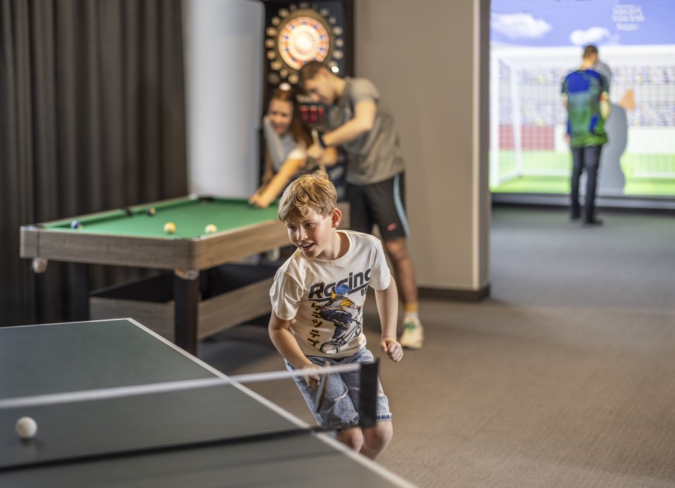 Un giovane ragazzo che gioca a ping-pong, con adolescenti che giocano a biliardo sullo sfondo.