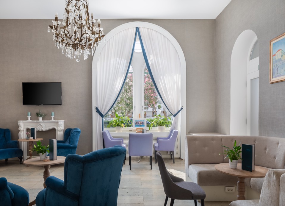 Interno del G&T lounge bar all'interno dell'Aminess Korcula Heritage Hotel, con un elegante arredamento in blu e grigio e una finestra che si affaccia sulla lussureggiante vegetazione mediterranea della pianta dell'oleandro.