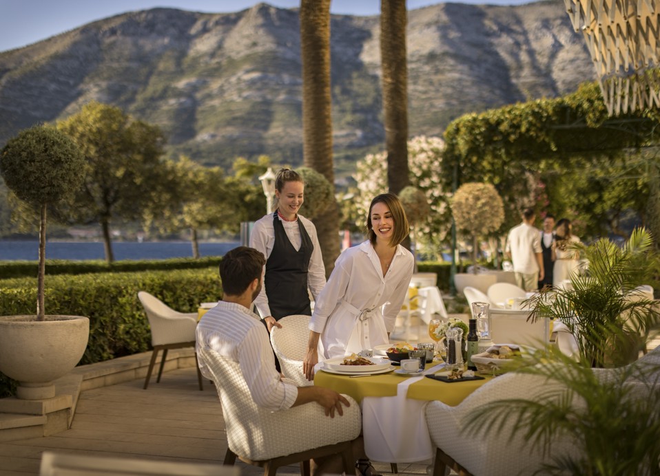Persone che cenano all'aperto al ristorante & bar a7 Seas con una vista panoramica su montagne e mare. Una donna in bianco si trova in piedi, sorridente, accanto a un uomo seduto. Un cameriere li serve.